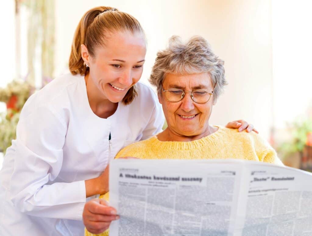 memory care worker helping older woman read newspaper